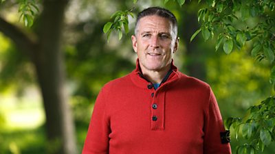 Iolo Williams outside with greenery behind him. He's wearing a red knitted jumper