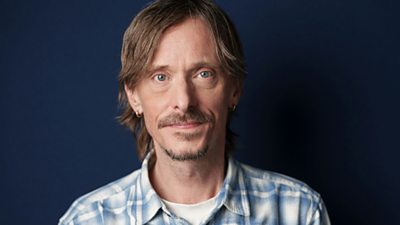 Headshot of Mackenzie Crook in front of a dark blue background.