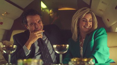 Tom Cullen and Stefanie Martini on set of The Gold in character as John and Marnie Palmer, sat smiling by a table with drinks in front of them