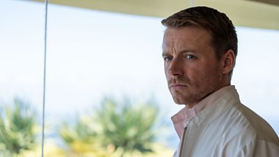 Jack Lowden on set of The Gold in character as Kenneth Noye. A man in a pale pink shirt stood next to a big glass window. It's sunny outside and there are palm trees