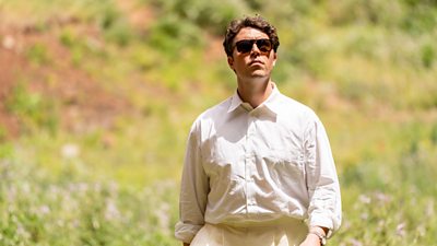 Tom Hughes on set of The Gold in character as Logan Campbell. He is stood in a field wearing a white shirt and sunglasses.