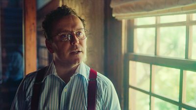 On set of The Gold, Joshua Mc Guire is in character as Douglas Baxter. He wears a stripe shirt with red braces and is stood next to a window.