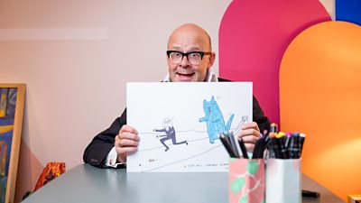 Harry Hill sat at a desk surrounded by art supplies and holding a drawing of someone running away from a big blue cat