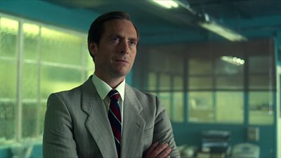 A man in a grey suit with his arms folded, looking across a room