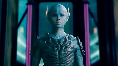 A blue fish like creature with human facial features stands looking solemn