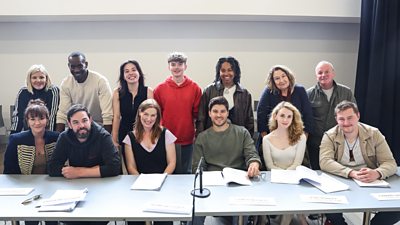 A group shot of thirteen cast members stood around a read-through table with open scripts laid out in front of them. They are all smiling.