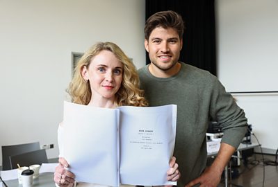 Jenn Murray and Cameron Cuffe stood together holding a paper script open . Behind them are some used coffee cups on a table
