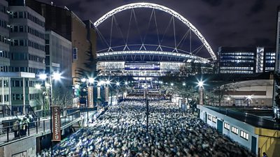 A shot of Wembley Stadium at night, taken from on Wembley Way, where thousands of spectators are on their way from an event there.
