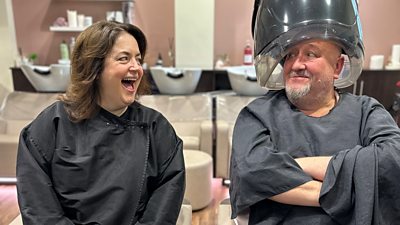 Photo from the production of From Merthyr with Love. Ruth Jones and Steve Speirs sit in a hair salon. Ruth laughs at Steve who sits under a large hairdryer and grins.