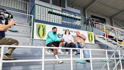 Photo from the production of From Merthyr with Love. Ruth Jones and Steve Speirs sit on bleachers at the Merthyr RFC stadium. They laugh together as they speak with a man in sportswear. A camera operator appears on the left of the image, filming their conversation.