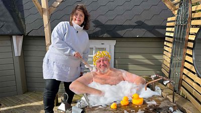 Photo from the production of From Merthyr with Love. Ruth Jones and Steve Speirs pose for the camera outdoors. Steve sits in an outdoor bubblebath beside three yellow rubber ducks. Ruth stands beside him in warm outdoor clothing, with a comical, panicked expression on her face.