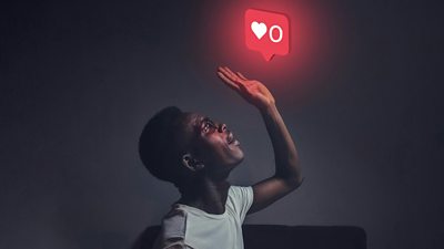 A person in a gloomy room looks up to and is illuminated by a red glowing social media 'love' icon hovering above. They are raising their hand - maybe to tap or hit the icon, maybe shielding themselves from its light.
