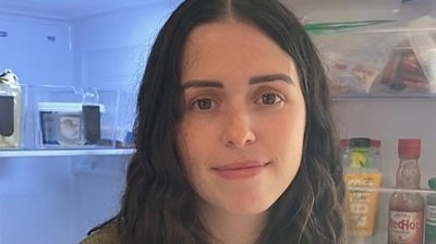 A woman with long dark hair looks into the camera. She has brown eyes and is standing in front of an open fridge.