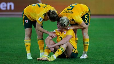 Two female footballers consoling another female footballer who is sat down on a football pitch looking upset. They are wearing yelow Wales away kits.