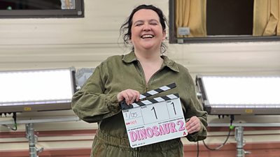 Ashley Storrie stands on the set of Dinosaur series two and holds a clapperboard. Pink, bubble writing on the board reads: “Dinosaur 2”.