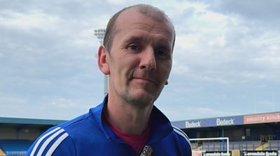 A man wearing a blue Glenavon zip up top with white stripes on the shoulders. He has light brown hair which is balding.