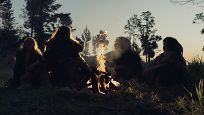 Four humans sat around a fire outside on a grassy patch of land. The image is dimly lit and so only their shadows are visible against the sky.