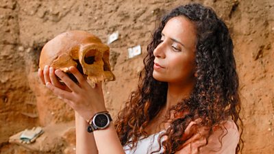 A woman holding a skull. She is looking at it closely.  Behind her the rocks are orange in what appears to be a dig site.
