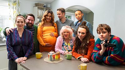 The eight cast members of Here We Go sat around a kitchen island with mugs of tea.One woman is pregnant and one of the boys is holding a video camera