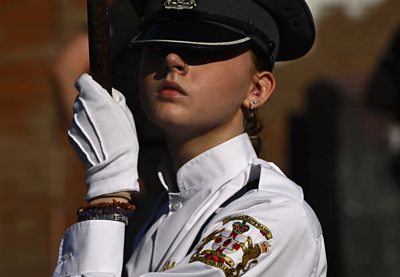 A young woman in a white shirt, white gloves and black peaked cap holds a flagpole