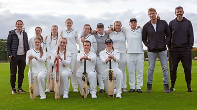 A young cricket team wearing white posing for a team photo on the field accompanied by three adults, one of which is Freddie Flintoff. The grass is very green and some of the team are holding cricket bats.