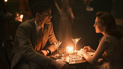 A young man and woman sat at a small round table, lit by candlelight drinking martinis. There is a glass ashtray on the table and a cigarette packet.