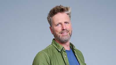A headshot of a man smiling against a grey background. He is smiling and wearing an open green shirt over a blue t-shirt. He also has a beard.