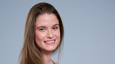 A headshot of a woman smiling against a grey background. She has long hair.