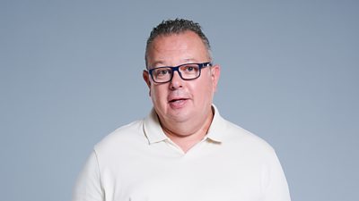 A headshot of a man against a grey background. He wears glasses and a white polo top.