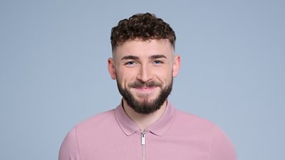 A headshot of a man smiling against a grey background. He is wearing a pink top with a zip. He has a beard.