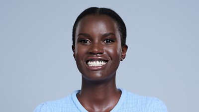 A headshot of a woman smiling against a grey background. She wears a baby blue top.