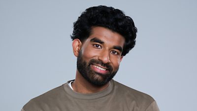 A headshot of a man smiling against a grey background. His head is tilted slightly to the side. He has a beard.