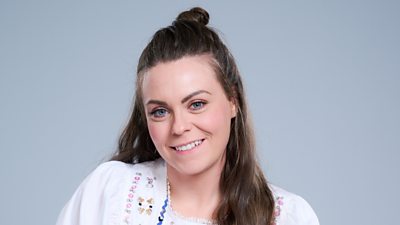 A headshot of a woman smiling against a grey background. She wears an embroidered white blouse.