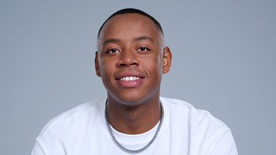 A headshot of a man smiling against a grey background. He wears a white t-shirt and a silver chain.