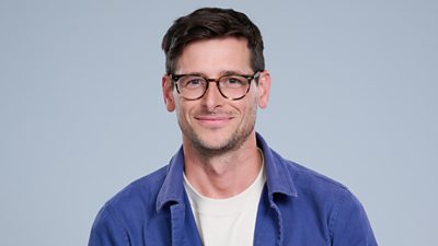 A headshot of a man smiling against a grey background. He wears an open blue shirt over a t-shirt.