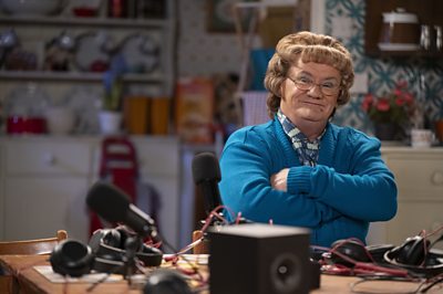 Mrs Brown sat at a wooden table in a house, wearing a blue cardigan with her arms folded. There are several pairs of headphones on the table.