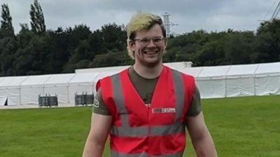 A man with blonde hair walks towards the camera wearing a pink high visibility jacket