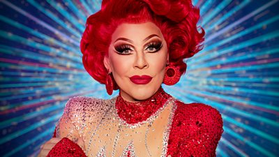 A head and shoulders shot of La Voix, a drag performer in a red wig and glittering red dress, with red earrings and a bold statement red lip.