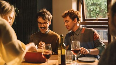 Characters sat around a dining table with plates, a serving dish, a wine bottle and glasses