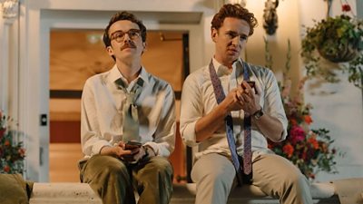 Alex Lawther and Laurie Kynaston sat in front of a white building with hanging baskets holding their mobile phones. They are wearing suits.