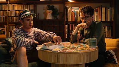 Two young men playing a board game around a small round table in a dark-ish room. There is a bookcase behind them and they look deep in thought.