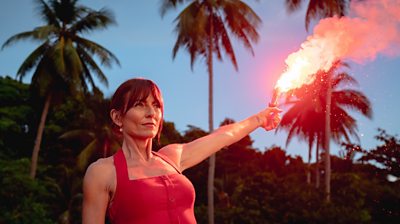 Close-up image of Davina McCall. Her arm is outstretched and holding a red flare. Tall palm trees are visible in the background. 