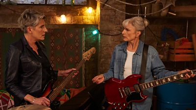 Two women holding guitars stand in a basement decorated with fairy lights. They look at each other with concerns expressions. A drum kit is visible in the corner of the image. 