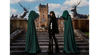 Claudia Winkleman stands on the steps of the Traitors castle, dressed in black with a black cape. Two hooded figures in green cloaks stand beside her. A castle and a staircase flanked by two statues of stags is visible behind them.