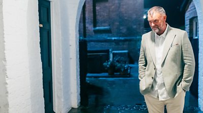 A man stood in a doorway.  He is wearing a linen jacket and pocket square.