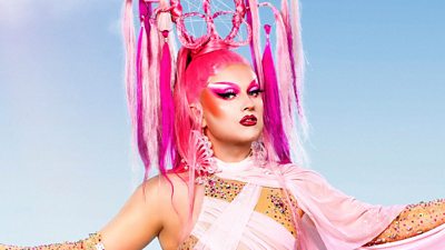 A drag queen wearing pink and gemstones against a blue background