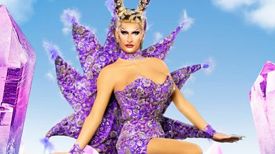 A drag queen wearing an elaborate purple outfit against a blue sky holding