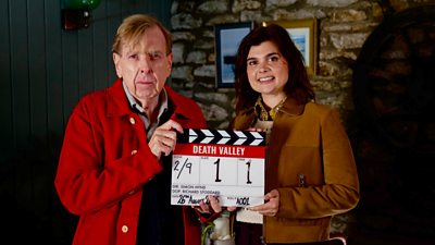 Timothy Spall and Gwyneth Keyworth holding a Death Valley clapperboard. They are stood in a dark room with exposed old brickwork.