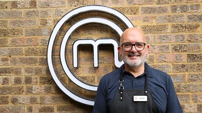 A portrait image of a contestant stood against an exposed brick wall, with the MasterChef circular white logo behind him.
