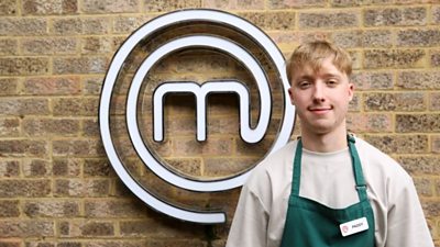 A portrait image of a contestant stood against an exposed brick wall, with the MasterChef circular white logo behind him.